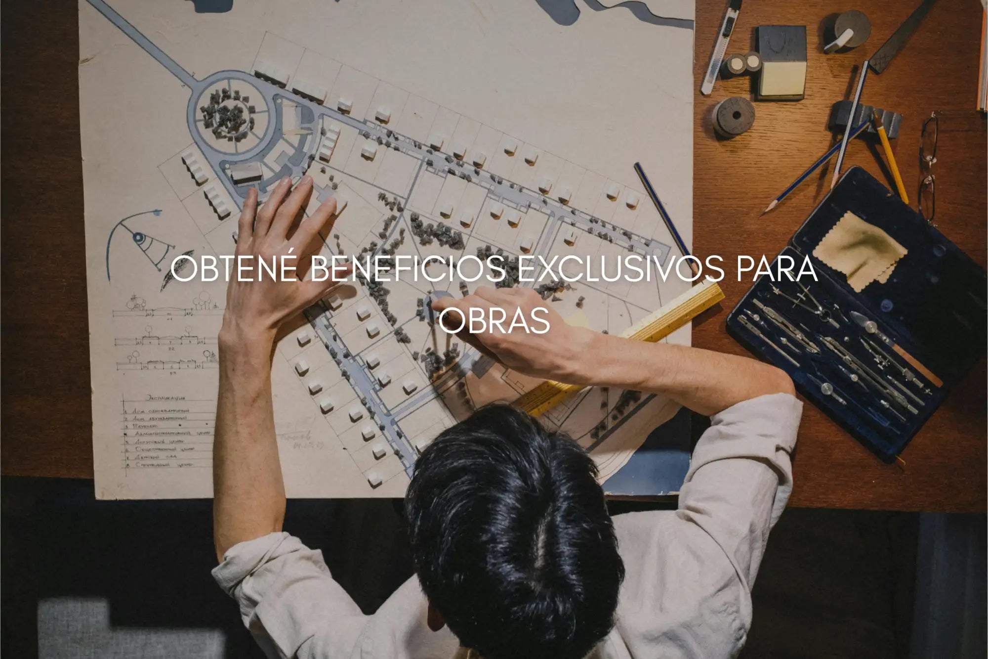 Una persona con una camisa de color claro trabaja en un escritorio de madera con planos arquitectónicos y herramientas de dibujo extendidas delante de ella.